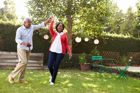 Mature couple dancing joyfully in a sunny backyard garden with green grass and hanging lanterns.