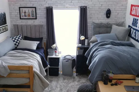 Cozy college dorm room with two beds, gray and blue bedding, a window with curtains, and personal decor.