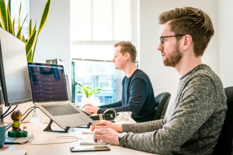 Two young men working on laptops and desktop computers in a bright modern office with plants