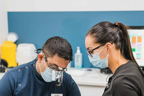 Dentist and assistant wearing masks and gloves performing dental treatment on patient in clinic.