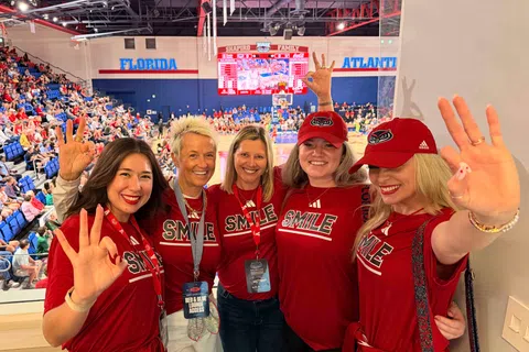 Five women in red SMILE shirts and hats smiling and making hand signs at a Florida Atlantic basketball game.