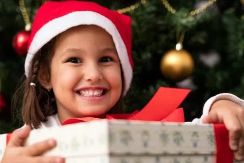 Smiling girl in Santa hat holding a wrapped Christmas present in front of a decorated tree with gold ornaments.