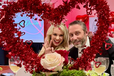 Smiling couple posing behind red heart-shaped decoration and roses at a romantic dinner event.