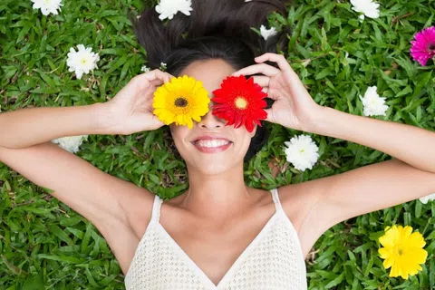 Smiling woman lying on grass holding yellow and red flowers over her eyes surrounded by white flowers.