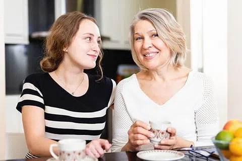 Two women sitting at a table drinking tea and smiling, with fruit bowl and snacks visible on the table.