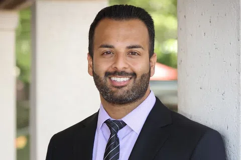 Smiling man in black suit and striped tie leaning against a wall with a blurred outdoor background.