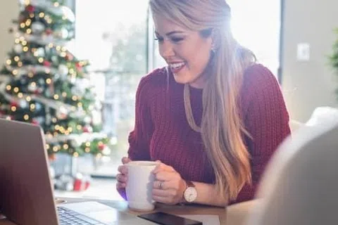 Woman in red sweater smiling while video chatting on laptop near decorated Christmas tree with holiday lights.