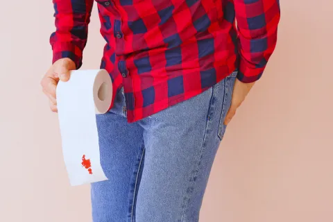 Person in red checkered shirt and blue jeans holding toilet paper with red stain against beige background.