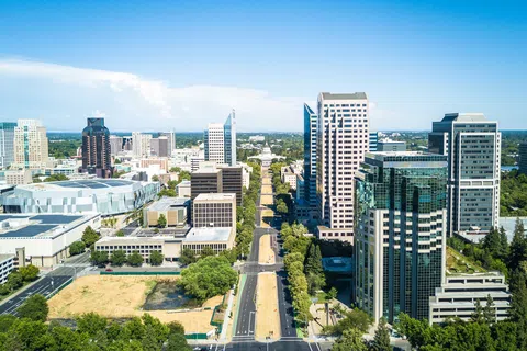 Aerial view of downtown Sacramento with skyscrapers, tree-lined streets, and clear blue sky.