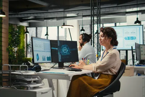 Woman working on AI and data analysis at a computer in a modern open office with colleagues and multiple monitors.
