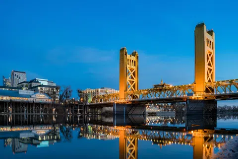 Golden Tower Bridge illuminated at dusk reflecting on calm river with city skyline in the background