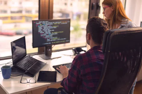 Two developers collaborate at a desk with multiple screens showing code in a modern office setting.