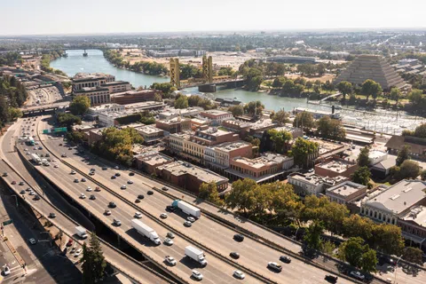 Aerial view of a busy highway and bridge over a river with urban buildings and trees on a sunny day.