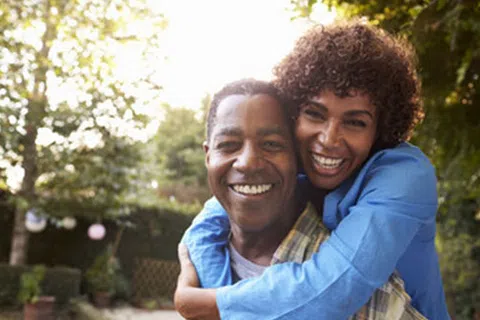 Smiling couple outdoors with woman hugging man from behind in a sunny green garden setting