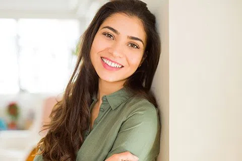 Smiling young woman with long dark hair wearing green shirt leaning against wall indoors.