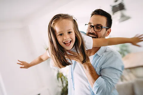 Father holding daughter flying indoors with arms outstretched, both smiling joyfully in a cozy home setting