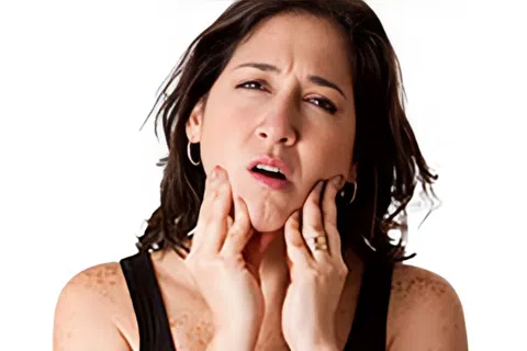 Woman in black tank top experiencing jaw pain holding her face with both hands against a white background