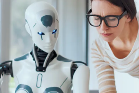 Woman in glasses leaning toward a robotic humanoid next to a computer in a bright office setting