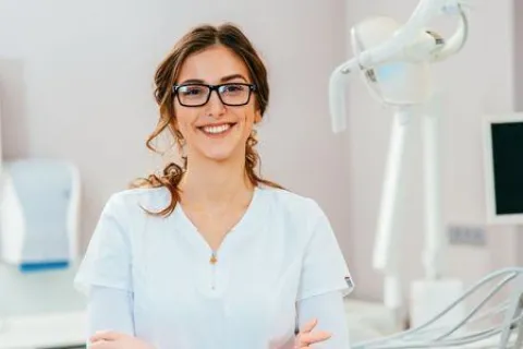 Smiling female dentist in white uniform standing in a modern dental clinic with equipment and monitor.