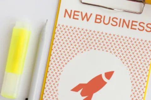 Clipboard with 'New Business' header, rocket graphic, pen, and yellow highlighter on a white desk.