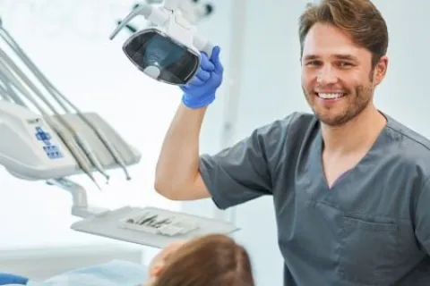 Smiling male dentist in gray scrubs holding dental light above patient in modern clinic
