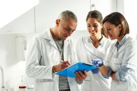Three medical professionals in lab coats discussing notes on a clipboard in a clinical setting with medical equipment.