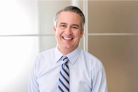 Smiling middle-aged man in light blue dress shirt and striped tie standing against office background.