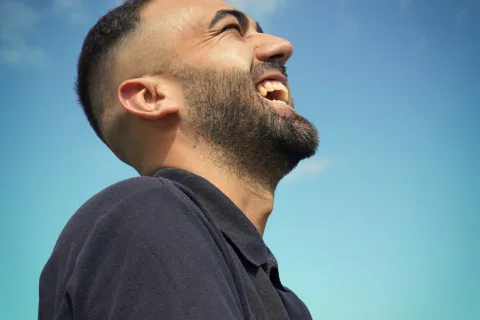 Side view of a bearded man laughing joyfully against a clear blue sky with a few clouds.