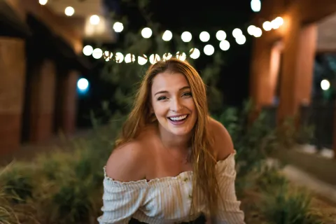 Smiling woman with long hair wearing an off-shoulder top outdoors at night with string lights in the background