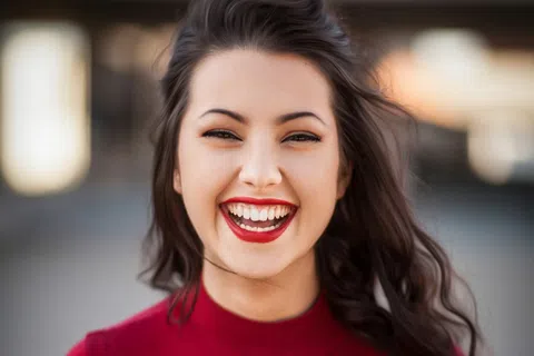 Young woman with dark hair wearing a red top smiling brightly outdoors on a blurred background.
