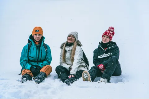 Three people sitting in the snow wearing winter jackets, hats, and gloves smiling at the camera outdoors.