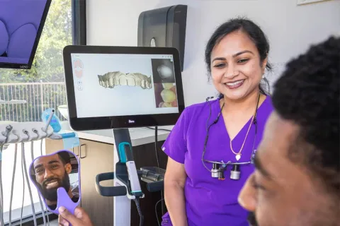 Dentist in purple scrubs smiling at patient holding mirror showing his teeth and dental 3D scan on screen