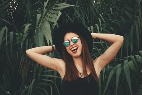 Smiling woman wearing a black hat and sunglasses stands with arms raised against lush green foliage.