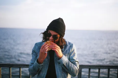 Person wearing sunglasses and a beanie eating a sandwich by a waterfront railing with calm water in the background
