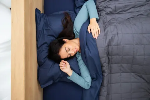 Woman sleeping peacefully in bed under a gray quilt and blue pillow, wearing a light blue long-sleeve shirt.