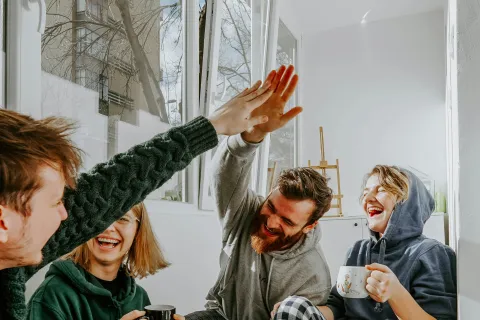Four friends in cozy clothes enjoying tea and laughing by a window, sharing a joyful high-five indoors.