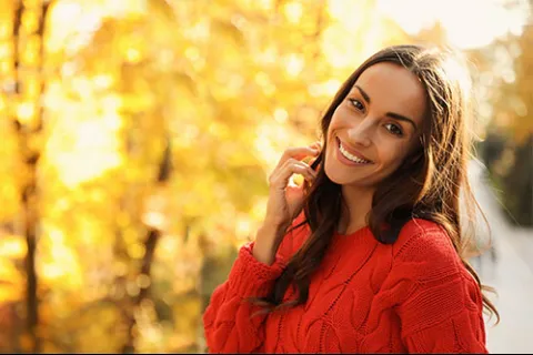 Smiling woman in a red sweater standing outdoors with blurred autumn foliage in the background