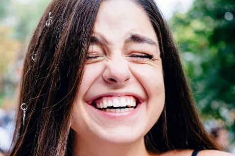 Young woman with long brown hair laughing joyfully outdoors wearing a black tank top with a blurred green background