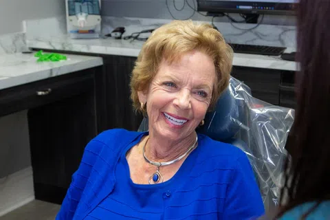 Smiling elderly woman in a blue top sitting in a dental chair during a consultation at the dental office.