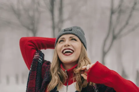 Smiling woman in a red cropped sweater, gray beanie, and plaid scarf enjoying a snowy winter day outdoors.