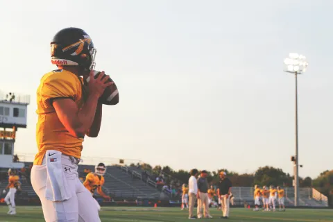 High school football player in yellow jersey prepares to throw ball during practice on field at sunset.