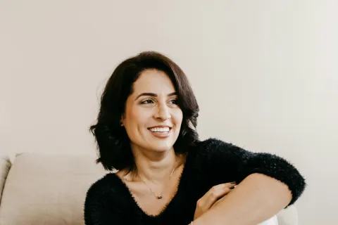 Smiling woman in black sweater and white pants sitting barefoot on striped couch indoors.