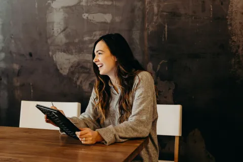 Smiling woman in gray sweater holding a tablet and pen sitting at a wooden table with dark textured wall background