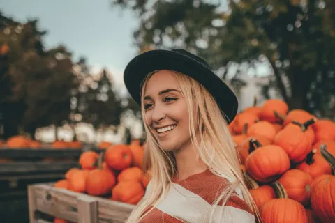 Smiling woman in striped sweater and hat holding a pumpkin by a wooden cart filled with pumpkins in autumn.