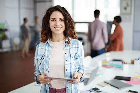 Smiling woman holding tablet in modern office with colleagues working in background