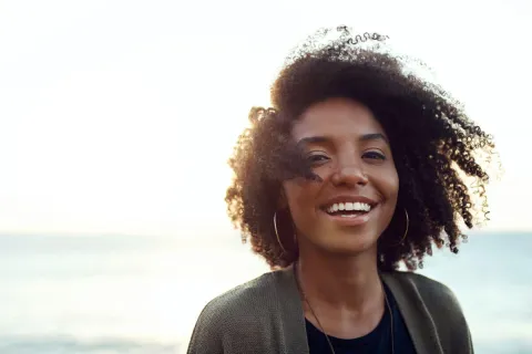 Smiling woman with curly hair against a beach sunset background, exuding warmth and happiness.