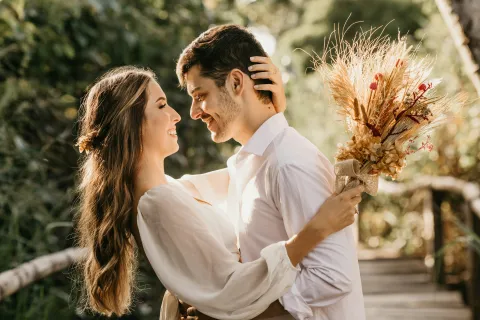 Couple in white clothing embracing outdoors with a dried flower bouquet on a sunny day.