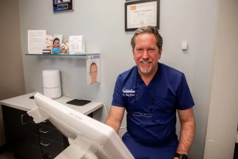 Smiling male doctor in blue scrubs sitting at desk with computer in a medical office setting.