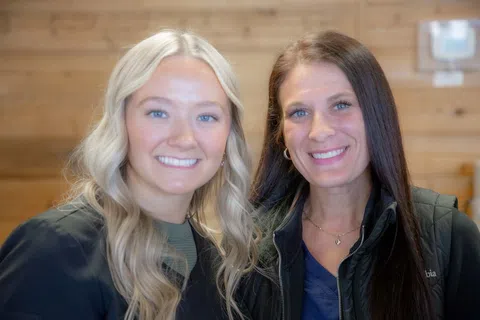 Two smiling women with blonde and brunette hair posing indoors in casual attire against wooden wall background
