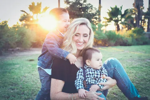 A joyful woman with two children, smiling in a sunny park surrounded by greenery and palm trees.